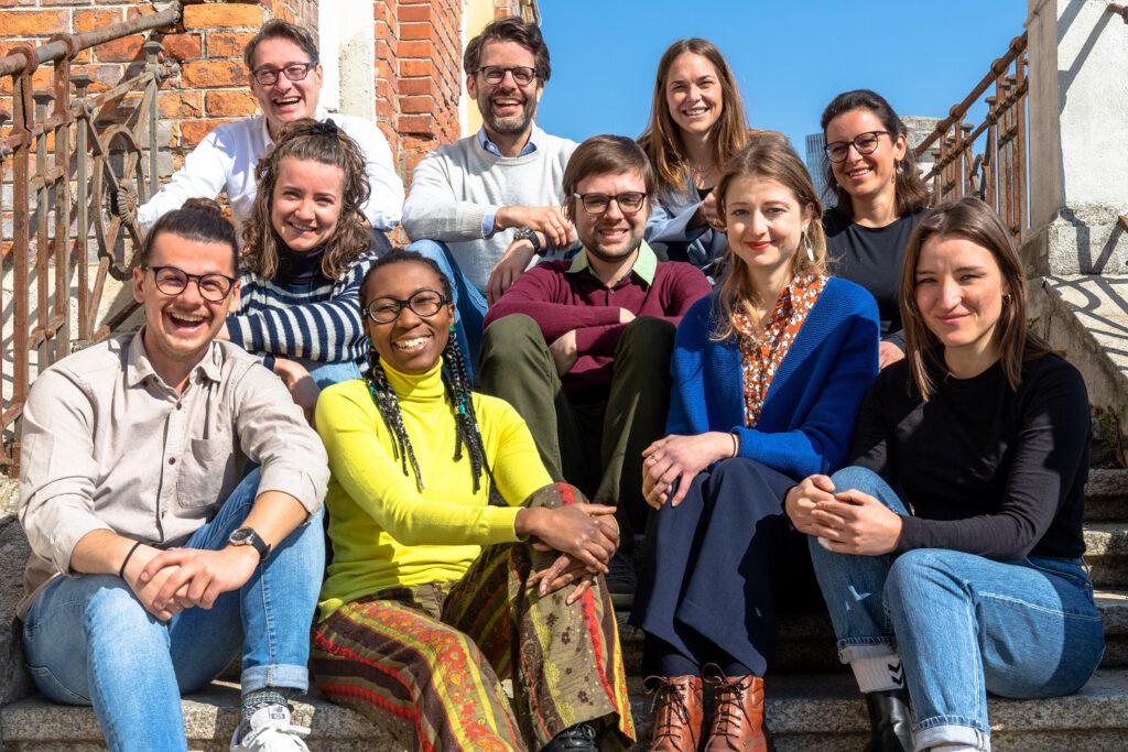 Die Trainees von Durchgang zwei sitzen gemeinsam mit dem Geschäftsführer, CCO und Team Lead des Traineeships für ein Gruppenfoto auf Stiegen.