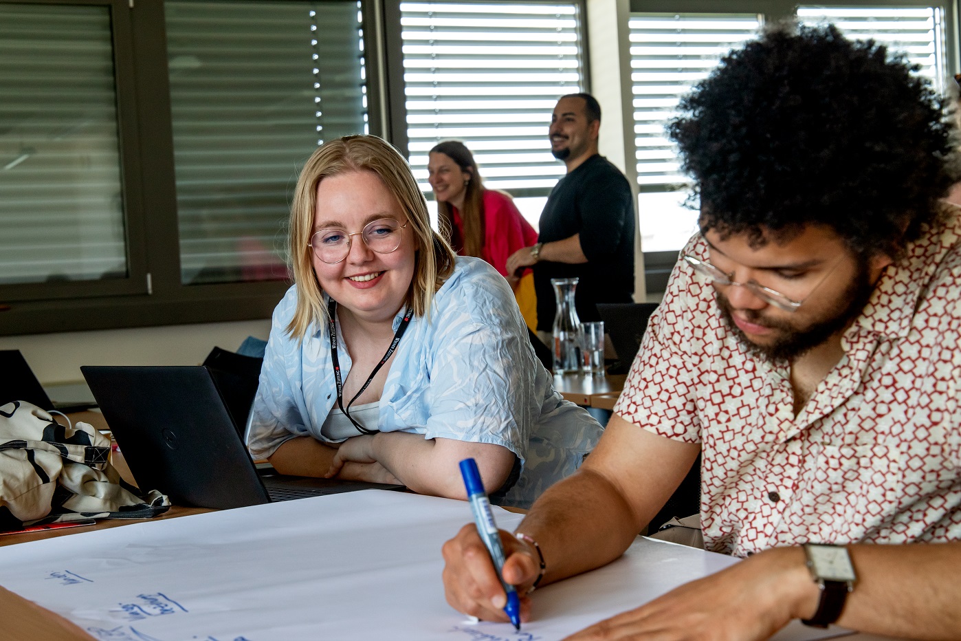 Ein männlicher Trainee schreibt mit einem Marker auf einem Flipchart Papier und seine Trainee Kollegin sagt ihm den Text an.